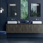Double vanity with two white sinks, dark wood cabinet, blue walls, two mirrors, and a plant-filled window between them.
