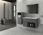 Modern grey bathroom featuring a glass shower enclosure, wall-mounted vanity, and a large mirror with slim light fixture above it. Beneath the mirror, open storage shows folded towels and a small display.
