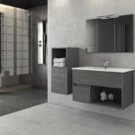 Modern grey bathroom featuring a glass shower enclosure, wall-mounted vanity, and a large mirror with slim light fixture above it. Beneath the mirror, open storage shows folded towels and a small display.