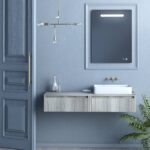 Blue bathroom with a floating wooden vanity, white rectangular sink, brass fixtures, and a large framed mirror panel; door on the left.