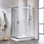 Frameless chrome-framed shower enclosure with sliding doors, rainfall showerhead, and handheld shower on the wall inside a modern bathroom; a striped towel hangs on the wall and a woven basket sits on the floor nearby.