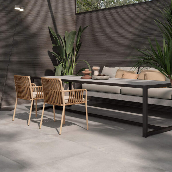 Modern outdoor dining area with a long table, wicker chairs, and neutral cushions under a glass skylight, surrounded by greenery.