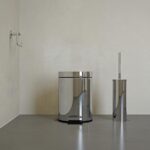 Polished chrome trash can with foot pedal and a matching toilet brush holder in a minimalist bathroom corner on a gray floor.