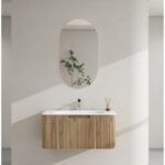 Modern bathroom with a curved white wall, an oval mirror, and a floating wooden slat vanity topped with a plant and diffuser reeds.