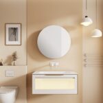Beige minimalist bathroom with a floating vanity, backlit drawer, large round mirror, pendant lights, framed art, a small vase, and a wall-mounted toilet.