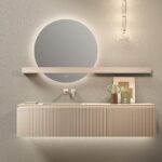 Round backlit mirror above a floating beige vanity with a slim wall shelf and pendant light in a minimalist bathroom scene.